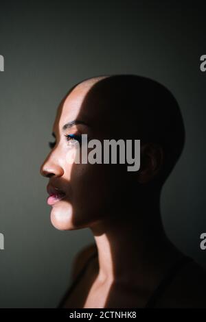 Vue latérale d'une femme afro-américaine attentionnés avec une coiffure chauve debout près de la fenêtre dans l'appartement et éclairé par sunbeam dedans matin Banque D'Images