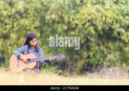 Une mignonne écolière asiatique aux cheveux longs jouait avec joie la guitare dans l'herbe le matin. Banque D'Images