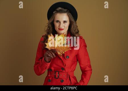 Bonjour novembre. Femme au foyer tendance de 40 ans, en robe rouge et béret noir avec gants en cuir et feuilles d'érable jaune d'automne isolées sur le backgro brun Banque D'Images