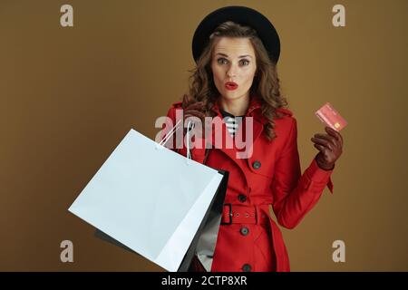 Bonjour octobre. Surprise élégante femme de 40 ans en manteau rouge et béret noir avec carte de crédit, gants en cuir et sacs de shopping en papier isolés sur b Banque D'Images