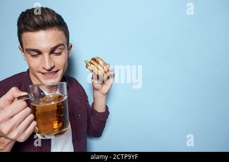 homme gai ivre avec une tasse de bière et un hamburger à la main régime alimentaire style de vie fond bleu Banque D'Images