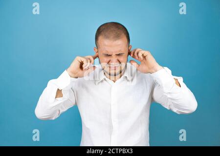 Homme d'affaires russe portant une chemise blanche sur fond bleu couvrant les oreilles avec les doigts avec une expression agacée pour le bruit de la musique forte. Banque D'Images