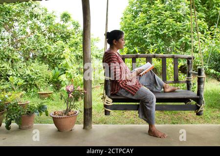 Une jolie femme assise sur une balançoire en bois lisant un réservez dans un environnement magnifique Banque D'Images