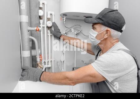 Le plombier mâle dans le masque médical vérifie les tuyaux pour le chauffage central et l'eau froide de l'appartement Banque D'Images