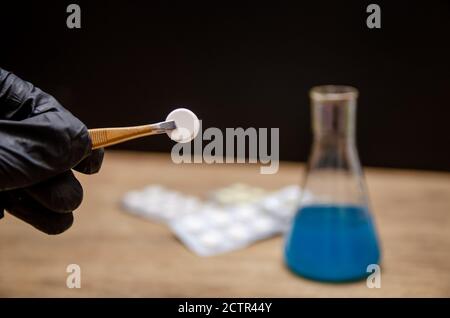 Une main en gants noirs tient des pinces et une pilule sur un fond flou de pilules et de flacons. Concept de soins de santé. Banque D'Images