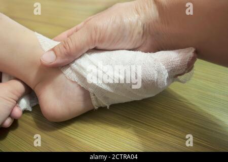 Une femme âgée de 35-50 ans bandage une plaie sur le pied d'un enfant de 5 ans. Le pied de bébé visible et les mains de la femme se rapprochent Banque D'Images