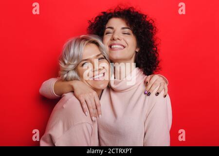 Les sœurs mignonnes aux cheveux bouclés dans les mêmes vêtements s'embrassent un mur rouge de studio Banque D'Images