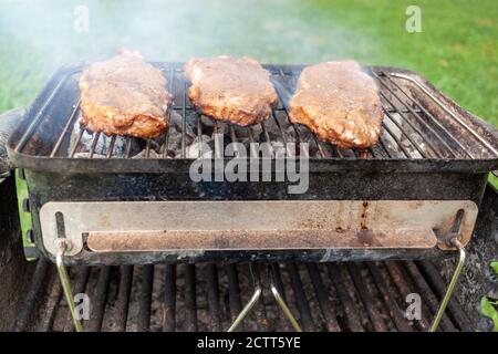 Gros plan image isolée de trois morceaux de steak de fer plat mariné sur un grill au charbon de bois. Le petit barbecue de pique-nique pliable est très chaud et de la fumée vient Banque D'Images