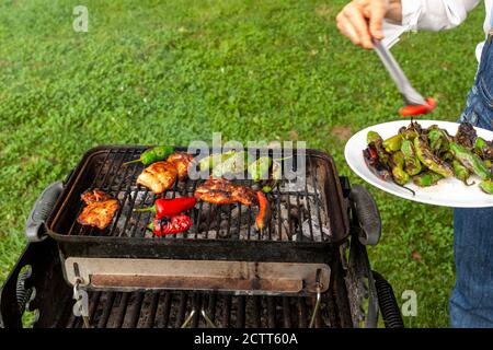 un choff portant jean combinaison et chemise blanche est par un grill au charbon de bois où les tranches de poulet marinées, les poivrons rouges et verts sont grillés au feu. Elle utilise Banque D'Images