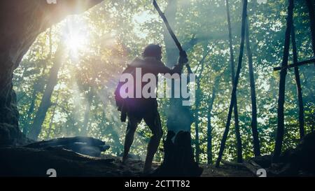 Le Caveman Primeval portant une peau d'animal tient une lance à pierre, se dresse à l'entrée de la grotte, donnant sur la forêt préhistorique prête à chasser les animaux préys Banque D'Images