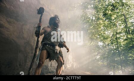 Primeval Caveman portant la peau d'animal tient le marteau de pierre près de Cave et regarde autour du paysage préhistorique, prêt à chasser la proie d'animal Banque D'Images