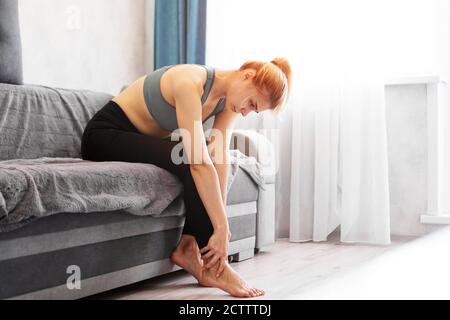 Une femme assise sur un canapé tient son pied blessé, se sentant mal. Concept de soins de santé et de médecine. Banque D'Images