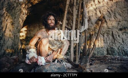 Primeval Caveman portant la peau d'animal tient Bone and Hits Rock avec elle. Neanderthal se promener près de l'entrée de la grotte, peut-être créer d'abord Banque D'Images