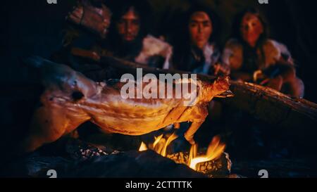 Gros plan de la famille Homo Sapiens cuisson de la viande animale au-dessus du feu de joie, puis alimentation. Tribu des chasseurs-cueilleurs préhistoriques portant des peaux d'animaux Banque D'Images