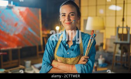 Portrait d'une jeune artiste féminine talentueuse sale avec de la peinture, portant un tablier, croise les bras tout en tenant des brosses, regarde la caméra avec un sourire Banque D'Images