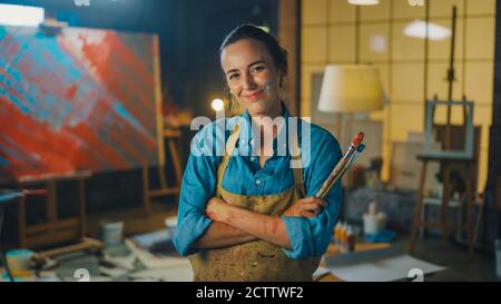 Portrait d'une jeune artiste féminine talentueuse sale avec de la peinture, portant un tablier, croise les bras tout en tenant des brosses, regarde la caméra avec un sourire Banque D'Images