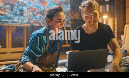 Deux artistes assis dans leur studio utilisent ordinateur portable, travailler sur un projet, parler et sourire. Painter et son partenaire faisant de la recherche Banque D'Images