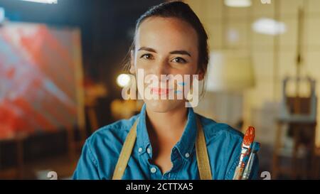 Jeune artiste femme professionnelle sale avec de la peinture, portant un tablier, bras croisés tout en tenant des brosses, regarde la caméra avec un sourire. Authentique Banque D'Images