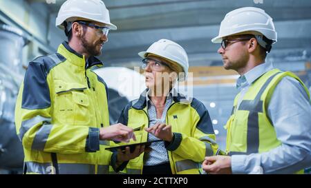 Trois ingénieurs de l'industrie lourde se trouvent dans l'usine de fabrication de tuyaux, utilisent l'ordinateur de tablette numérique, ont la discussion. Grand tuyau assemblé. Conception et Banque D'Images