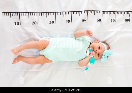 Un bébé de trois mois en vêtements vert menthe allongé sur un lit sur lequel est tiré une règle de mesure pour la croissance. Poussée dentaire Banque D'Images