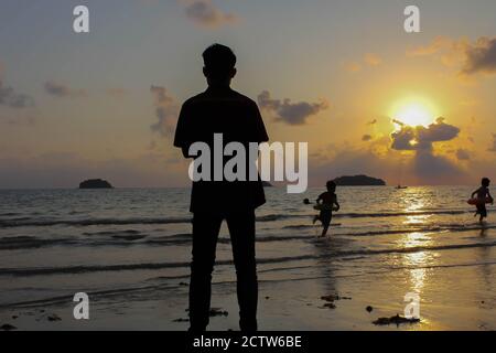 Homme solitaire se tient sur la plage regarde vers la lumière du soleil, marches sur le sable, coucher de soleil, silhouette de personne marchant sur la plage de mer. Banque D'Images