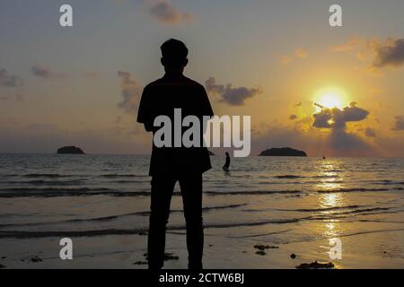 Homme solitaire se tient sur la plage regarde vers la lumière du soleil, marches sur le sable, coucher de soleil, silhouette de personne marchant sur la plage de mer. Banque D'Images