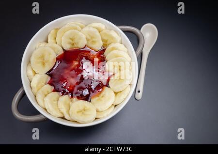 Flocons d'avoine aux bananes, confiture dans un bol gris-blanc avec une cuillère blanche sur fond noir, vue de dessus, espace de copie. Petit déjeuner sain. Banque D'Images