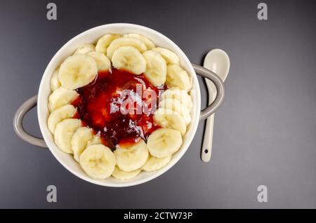 Flocons d'avoine aux bananes, confiture dans un bol gris-blanc avec une cuillère blanche sur fond noir, vue de dessus, espace de copie. Petit déjeuner sain. Banque D'Images