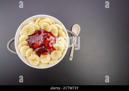 Flocons d'avoine aux bananes, confiture dans un bol gris-blanc avec une cuillère blanche sur fond noir, vue de dessus, espace de copie. Petit déjeuner sain. Banque D'Images
