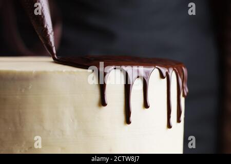 Le processus de décoration du gâteau avec du chocolat liquide. Un confiseur presse le chocolat liquide d'un sac de pâtisserie à un gâteau au biscuit à la crème blanche. Banque D'Images