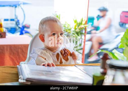 l'enfant regarde l'avant avec soin. Une jeune fille est assise sur une chaise haute de bébé dans un café de rue. Banque D'Images