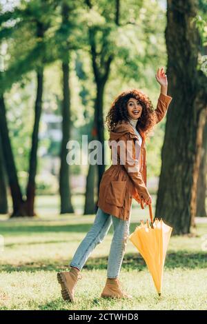 Vue latérale de la femme maurique avec un parapluie qui agite la main caméra pendant que vous marchez dans le parc Banque D'Images