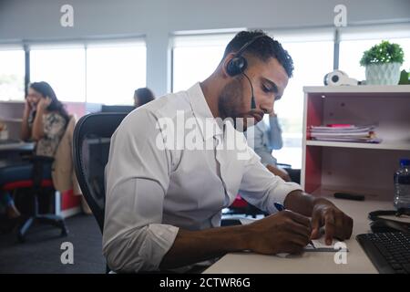 Élégant homme d'affaires de course mixte vêtu assis au bureau, portant un casque téléphonique et écrivant des notes, avec des collègues en arrière-plan. Bus créatif Banque D'Images