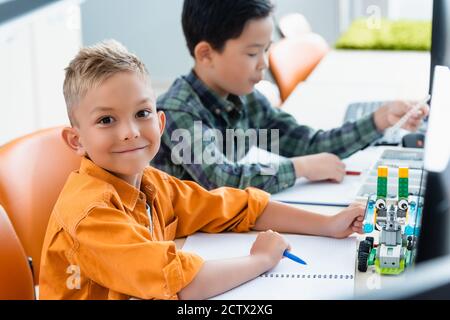 Mise au point sélective d'un élève regardant l'appareil photo près du robot et ami asiatique dans l'école stem Banque D'Images