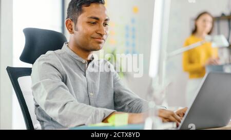 Un travailleur de bureau indien intelligent et élégant assis à son bureau travaille sur un ordinateur portable. En arrière-plan Bureau moderne avec équipe diversifiée de jeunes Banque D'Images