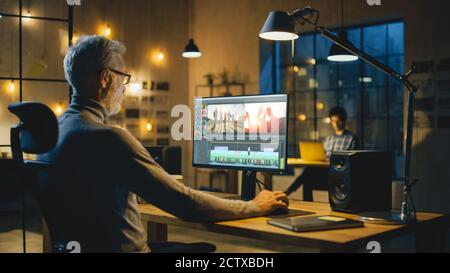 Soirée dans un bureau créatif : un vidéaste professionnel d'âge moyen travaille sur un ordinateur de bureau, fait du montage vidéo, à l'aide d'un logiciel d'édition vidéo Banque D'Images