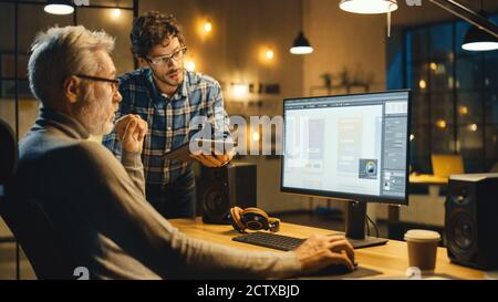 Le soir, Creative Middle Aged Mobile Software Designer utilise un ordinateur de bureau avec smartphone application Design, a la conversation avec plus jeune Banque D'Images