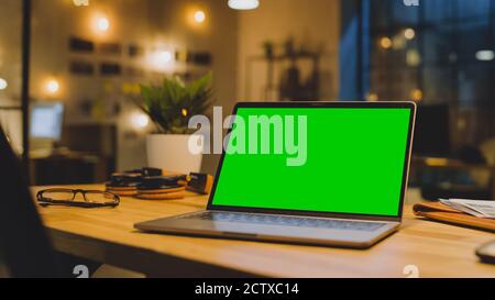 Maquette d'écran vert Stanting sur le bureau. En arrière-plan élégant studio moderne de bureau dans la soirée avec grande fenêtre CityScape Banque D'Images