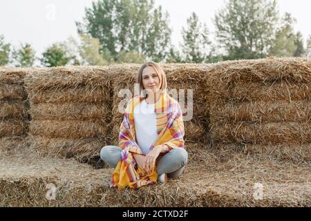 Portrait de femme automne. Portrait d'une belle femme d'âge moyen du Caucase assise sur une haystack à la ferme. Femme pensive enveloppée d'une écharpe jaune Banque D'Images