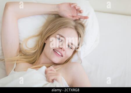 Belle jeune femme blonde couchée dans le lit souriant à la caméra. Banque D'Images
