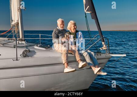 Célébration de l'anniversaire de mariage. Joyeux couple senior qui boit du vin ou du champagne et rit tout en s'asseyant sur un voilier ou un pont de yacht flottant dans un Banque D'Images