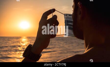 Portrait de l'homme dans un masque médical sur son visage debout sur une belle plage avec lumière orange de coucher de soleil. Homme adulte avec un masque médical pour se protéger fro Banque D'Images