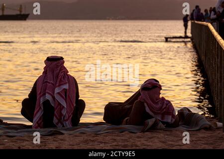 Deux hommes arabes portant du keffiyeh, agal et thobe sont vus sur la plage à côté d'un quai. L'un est allongé sur son dos, l'autre est assis à pattes croisées. Ils Banque D'Images