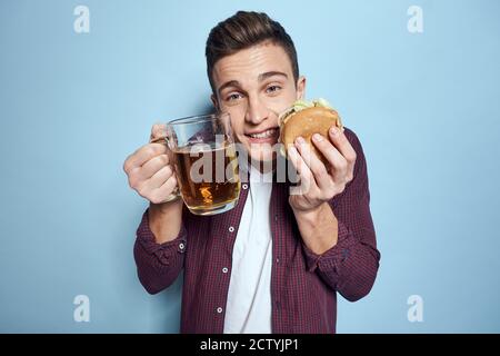 homme gai ivre avec une tasse de bière et un hamburger à la main régime alimentaire style de vie fond bleu Banque D'Images