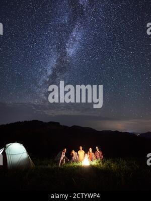 Camping de nuit. Groupe vertical de touristes ayant un repos autour de feu de camp dans les montagnes en été, ayant de petites discussions près de tente illuminée sous beau ciel plein d'étoiles et de voie lactée Banque D'Images