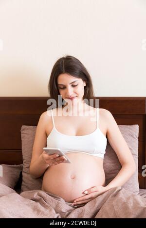 Femme enceinte caucasienne utilisant un téléphone portable, se reposant sur un lit à la maison. Banque D'Images