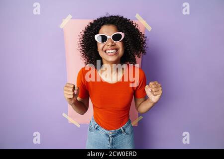 Image d'une femme afro-américaine excitée en lunettes de soleil faisant le gagnant geste isolé sur un arrière-plan multicolore Banque D'Images