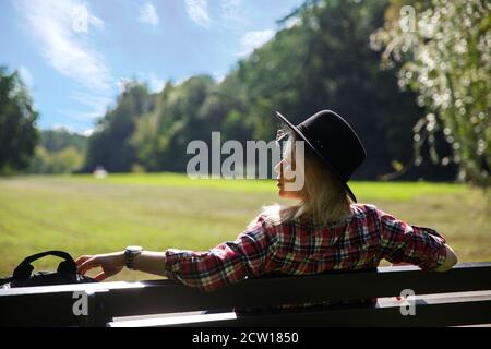 une jeune femme sous un chapeau et une chemise à carreaux est assise sur un banc près de la prairie. Banque D'Images