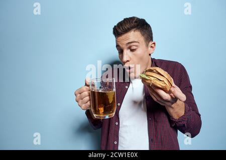 homme gai ivre avec une tasse de bière et un hamburger à la main régime alimentaire style de vie fond bleu Banque D'Images