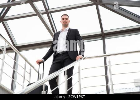 Jeune homme d'affaires portant un costume habillé debout sur un escalier à l'intérieur bâtiment moderne avec valise Banque D'Images